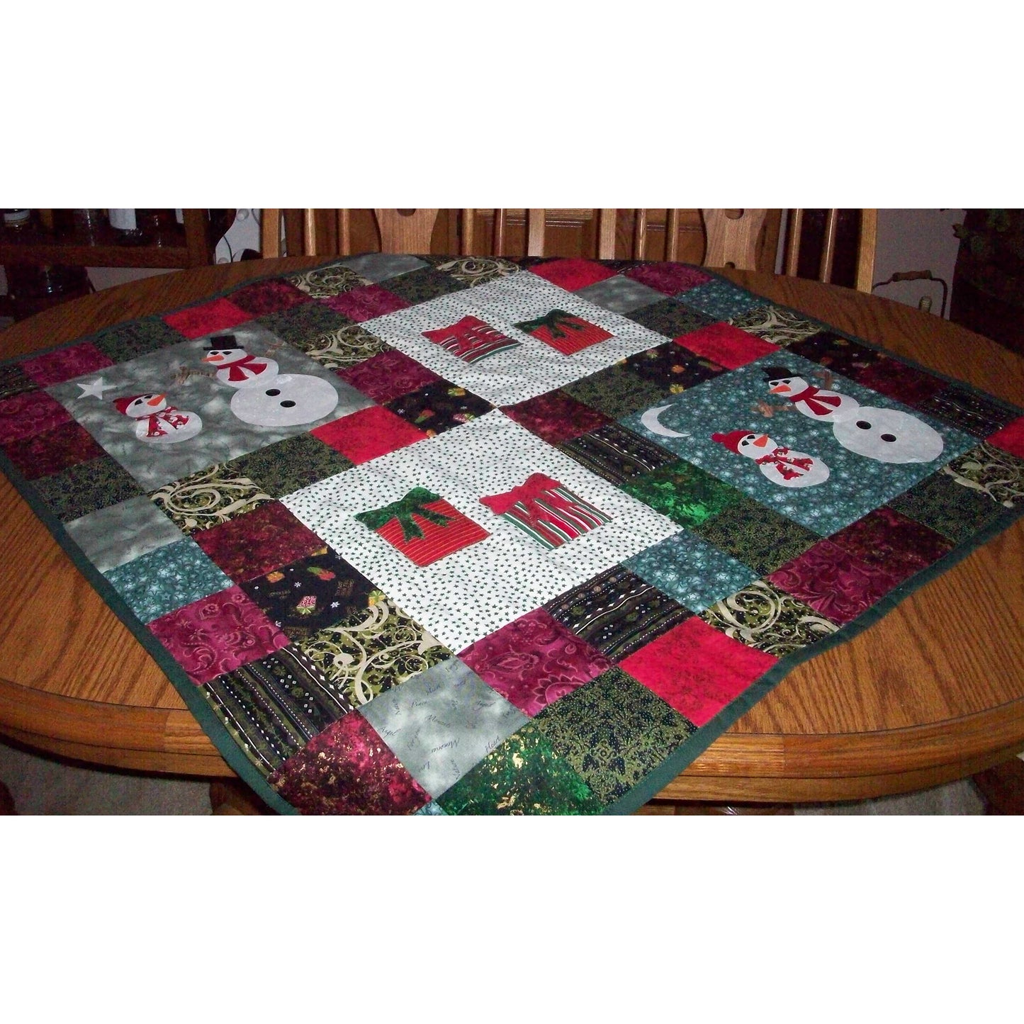 Adorable table topper pictured on a table. The table topper includes four blocks with snowmen in two and presents in the other two. The rest is scrappy with holiday fabric.