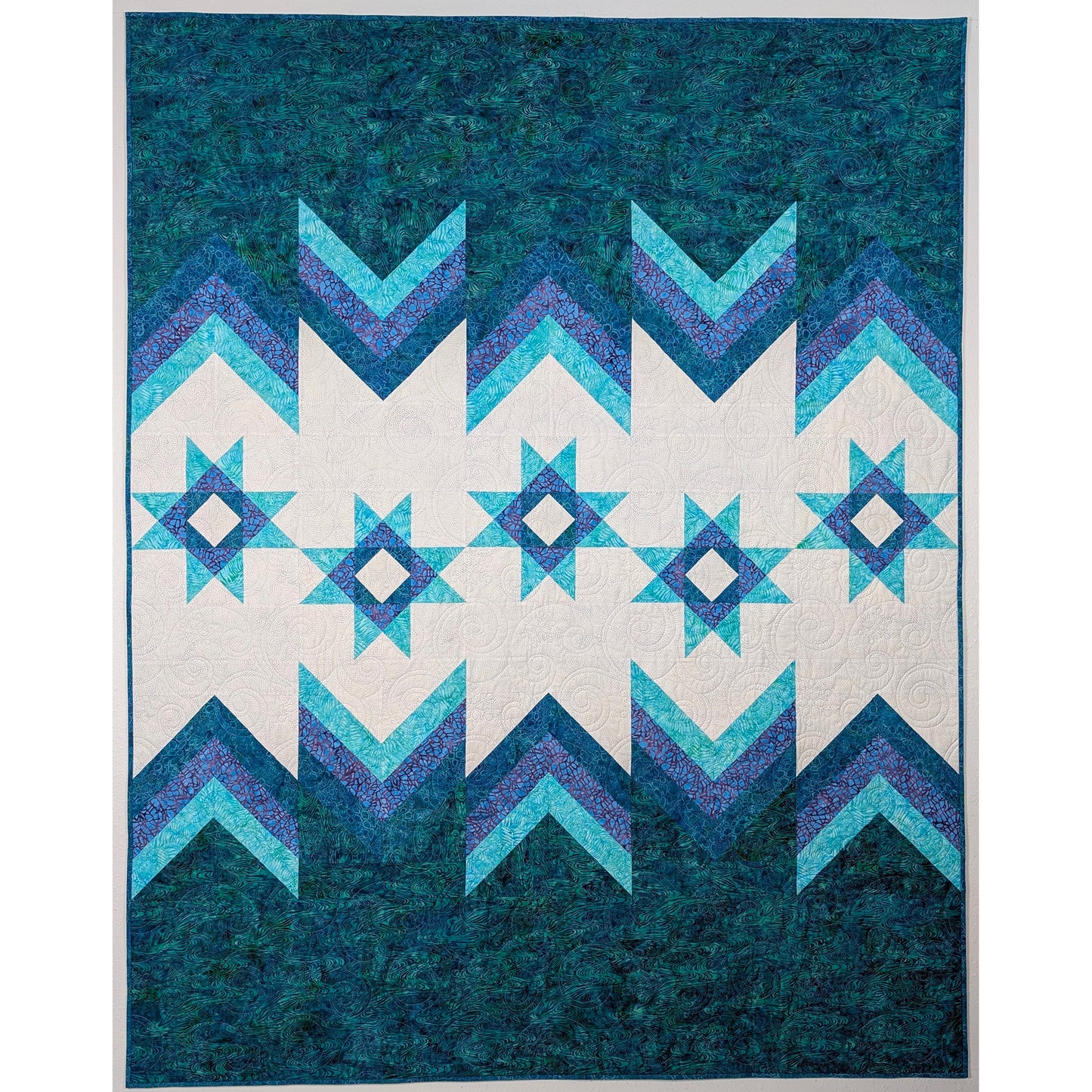 Beautiful quilt features a green bottom with variegated heights for either V or inverted V shapes of three shades of blue. Above this in a white background are Ohio Stars featuring a white center triangle outside a dark blue triangle. Light blue finish off the star points. Top is a similar variegated pattern but inverted.