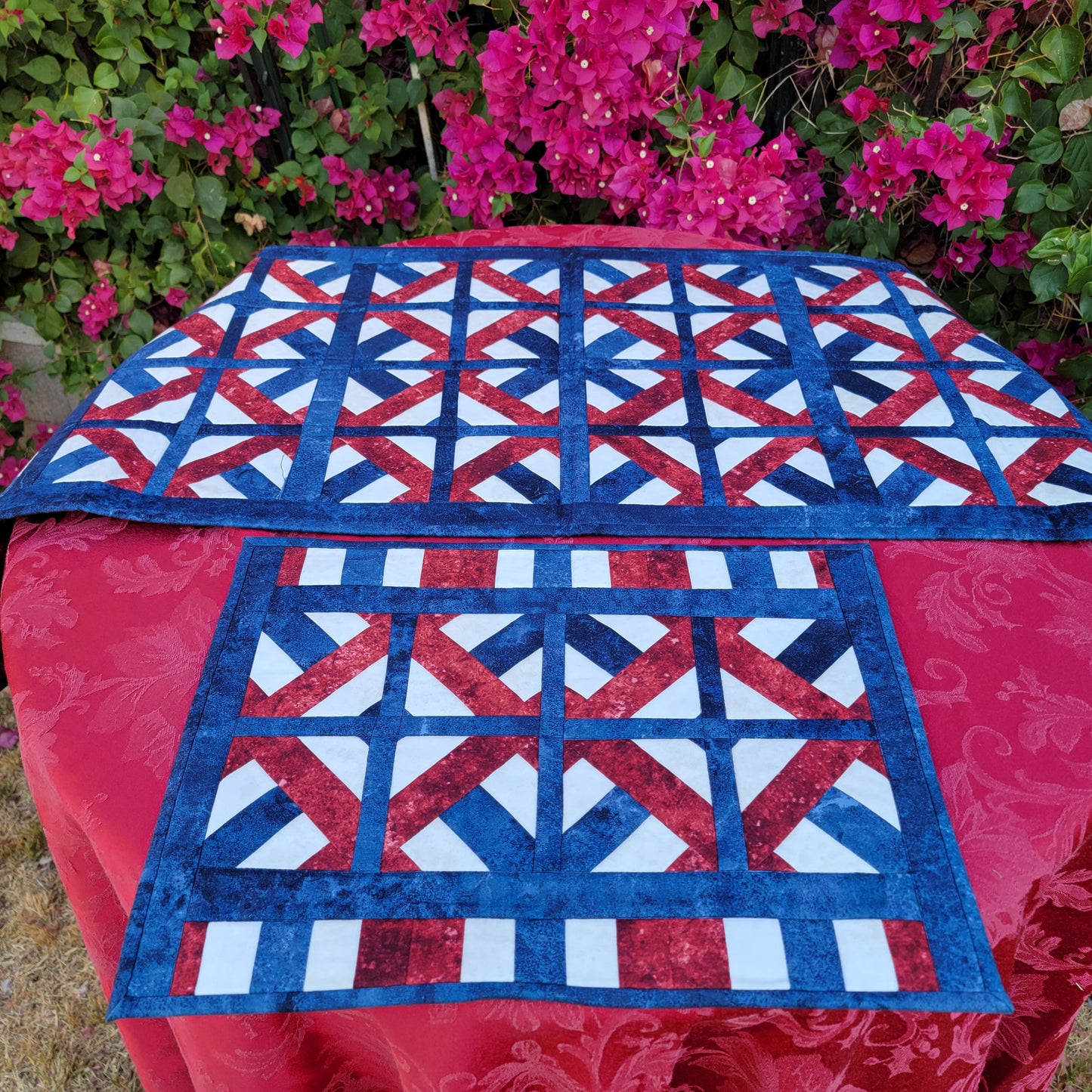 Quilt with red, white, and blue pattern on a table outdoors with pink flowers in the background