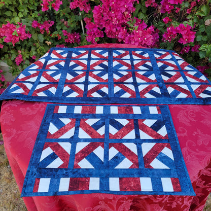 Quilt with red, white, and blue pattern on a table outdoors with pink flowers in the background