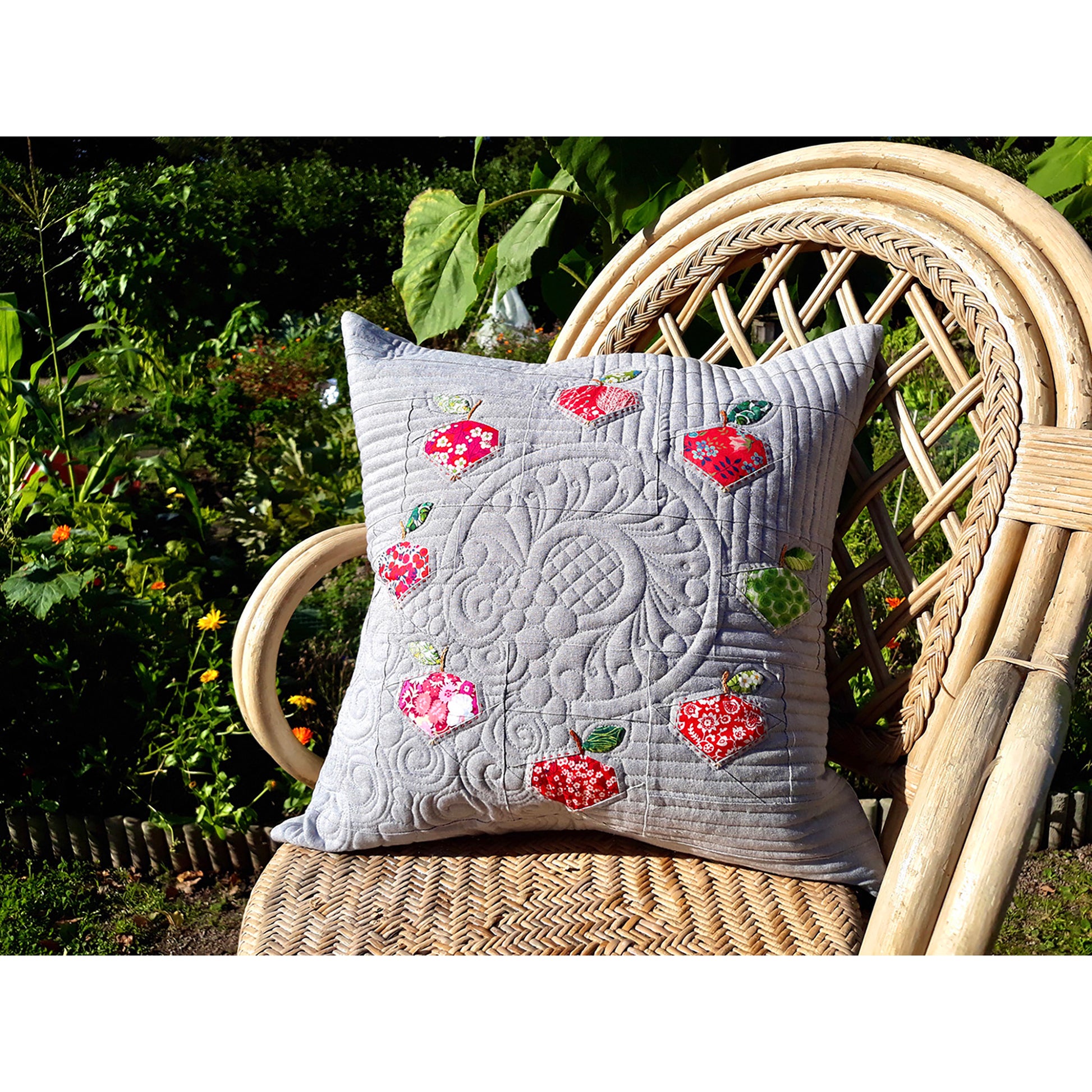 Beautifully quilted grey pillow with apples sitting on a wicker chair outside.