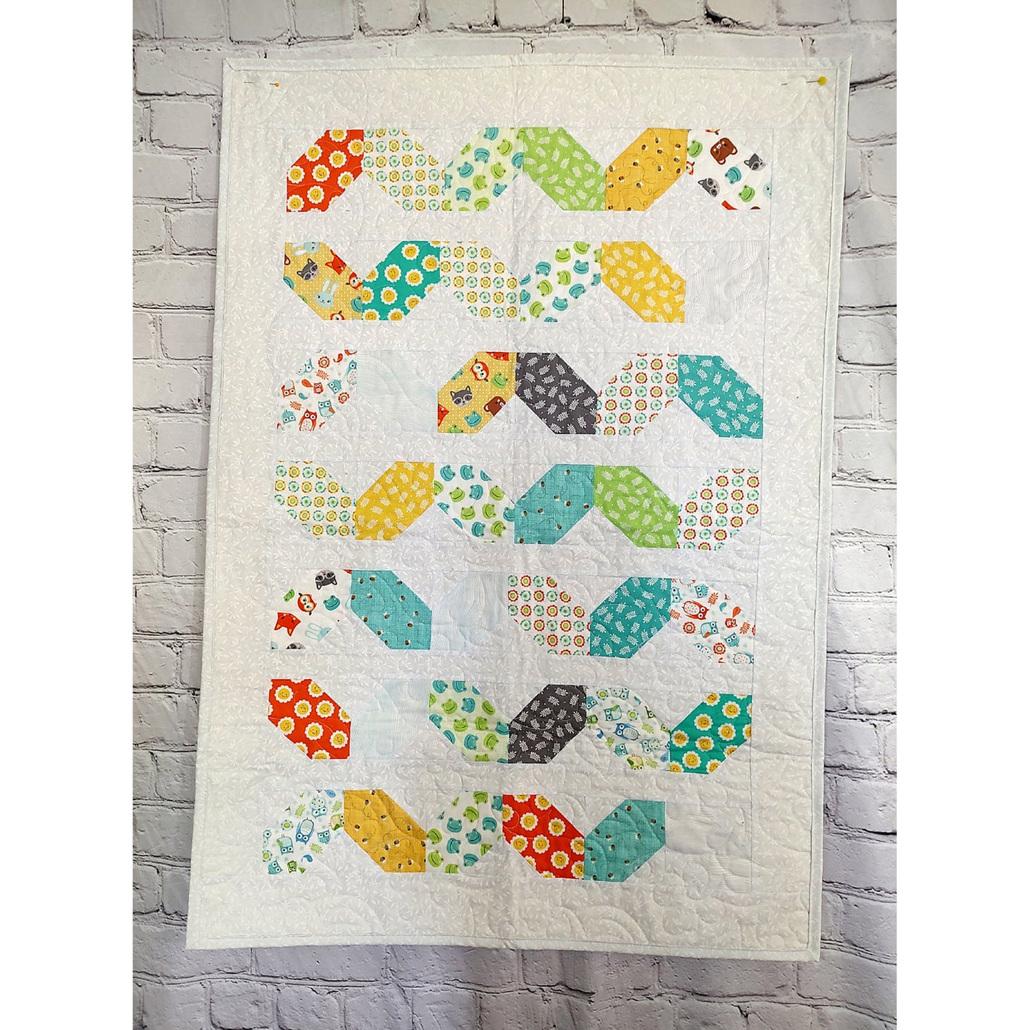 Bright quilt featuring a geometric pattern in various colors looking almost like a back and forth worm, colorful child's toy, or colorful baguettes.