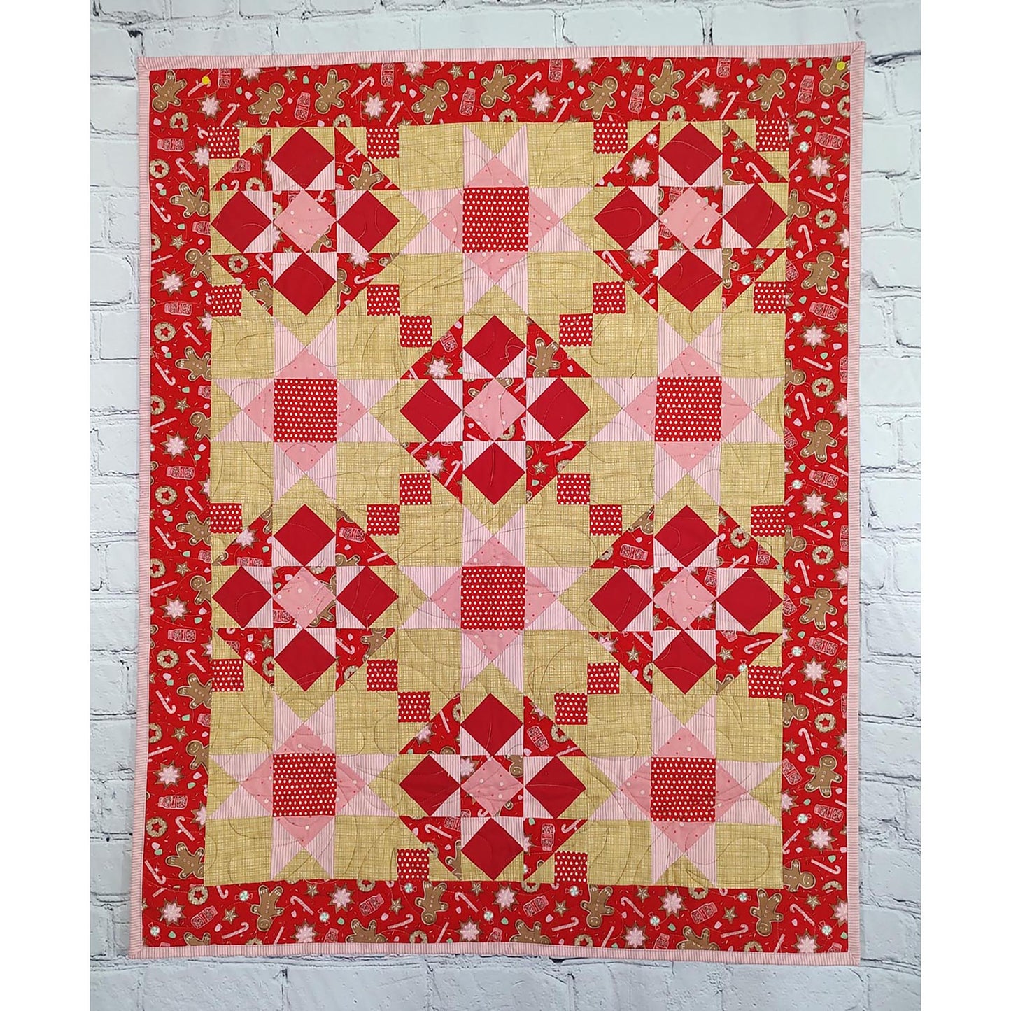 Festive quilt features red fabric with cookies and candy canes. Pieced into blasts of stars and diamond shapes with pink and cream fabrics added.