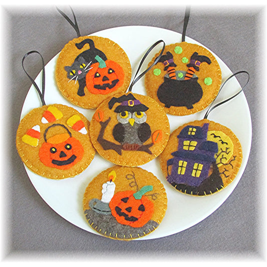 Plate full of adorable wool felt Halloween ornaments. 6 ornaments which include a black cat and jack o-lantern, a haunted house, a jack o-lantern next to a lit candle, an owl with witch's hat on a branch, a caldron with witch's feet sticking out, and a jack o-lantern candy basket with candy corn.