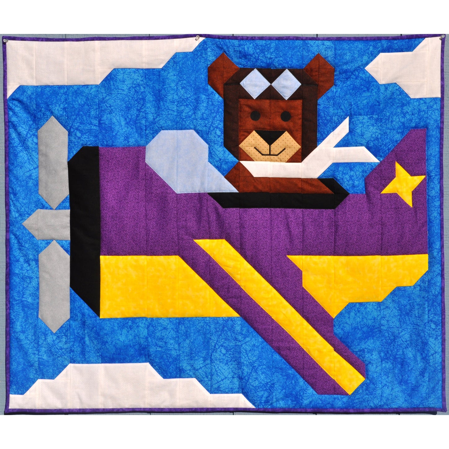Adorable quilt features a bear flying an airplane in a blue and slightly cloudy sky.