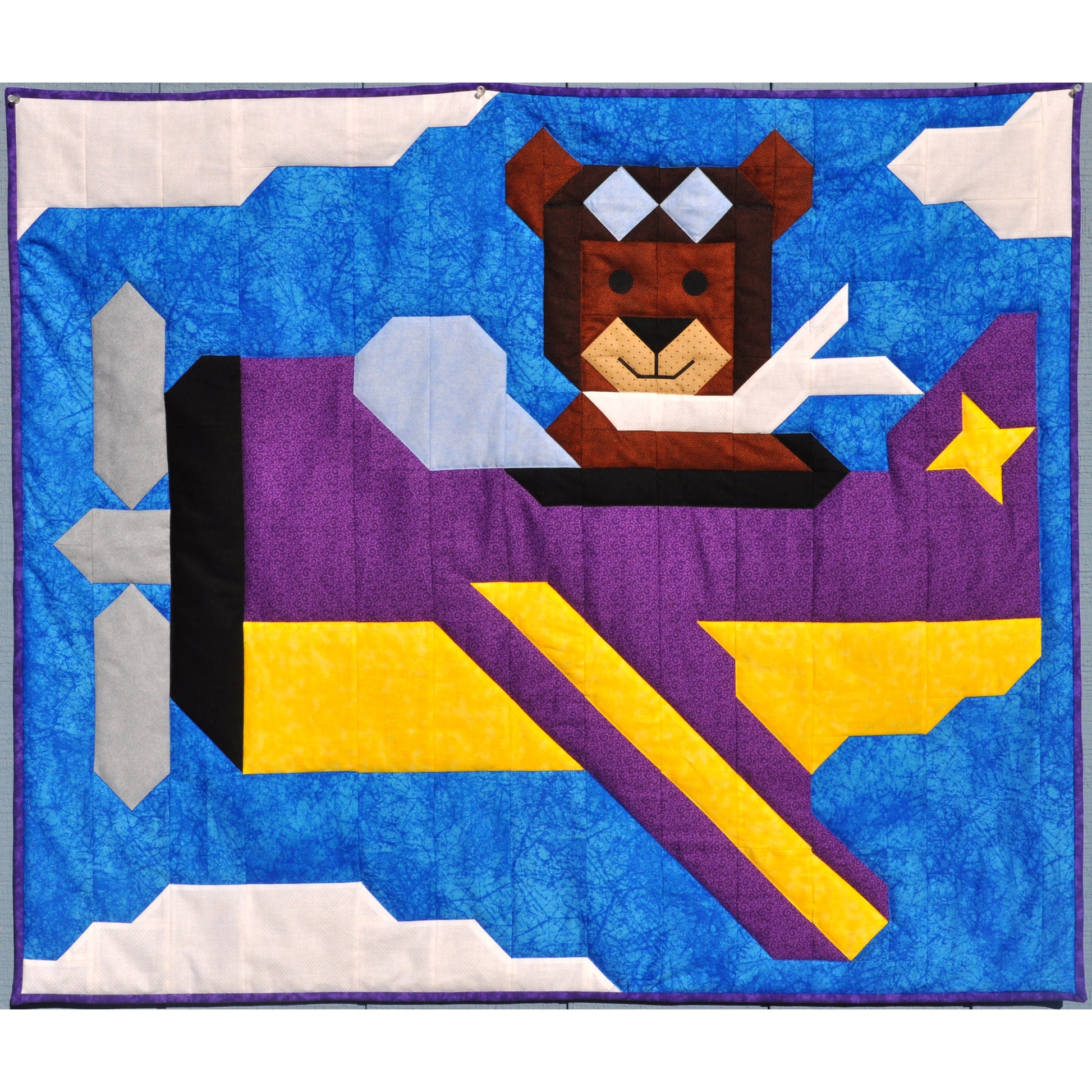 Adorable quilt features a bear flying an airplane in a blue and slightly cloudy sky.