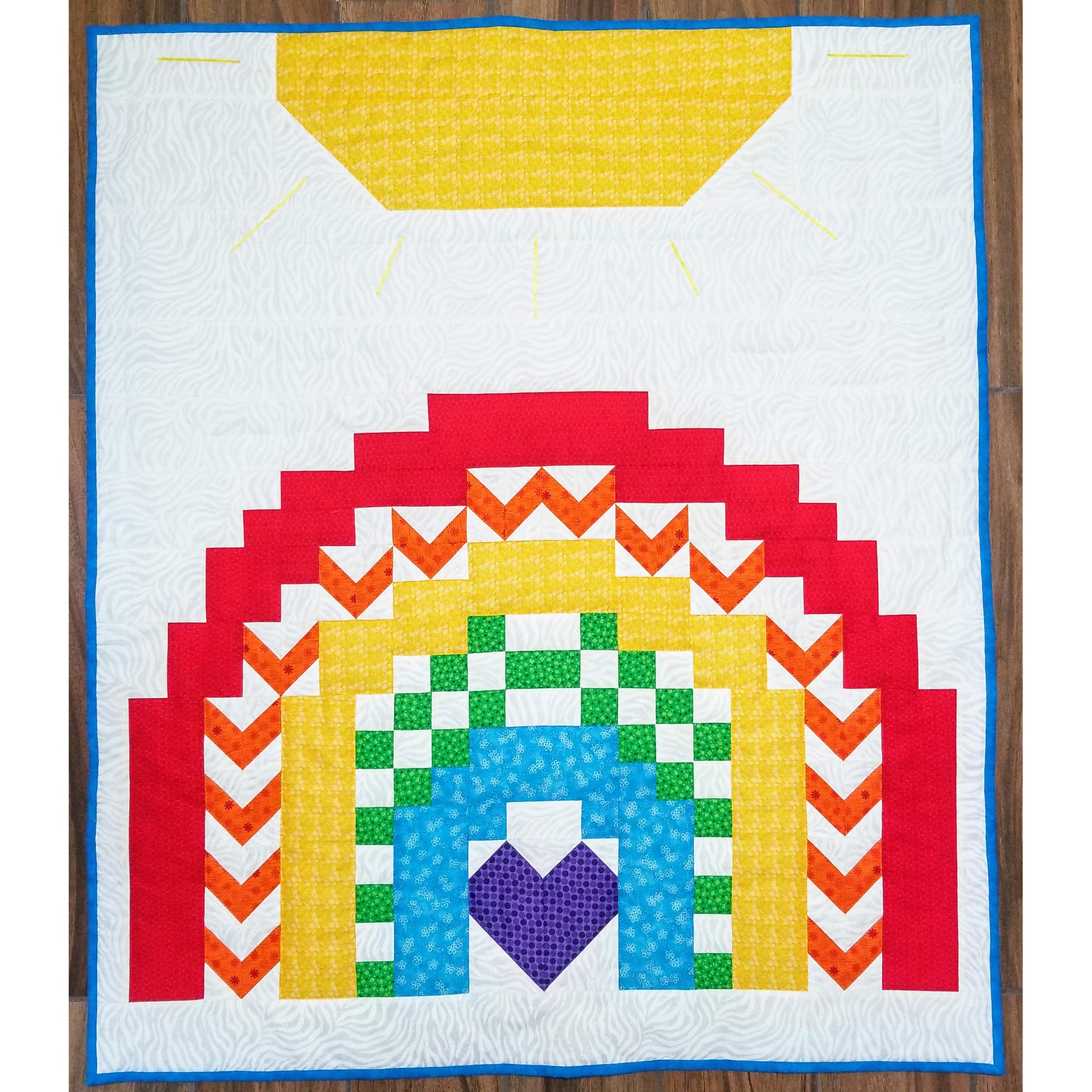 Rainbow quilt features a sun on the top with a pretty rainbow below. Each rainbow color has a different pattern from solid to V or blocks. The middle violet is in the shape of a heart.