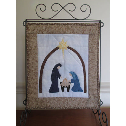 Festive quilt handing from metal stand. The quilt is the nativity scene with arch holding up the star in the middle. Below is a standing figure (probably Joseph), baby Jesus in manger, and kneeling Mary. Design in the middle has white background. Entire scene is framed by brown fabric.