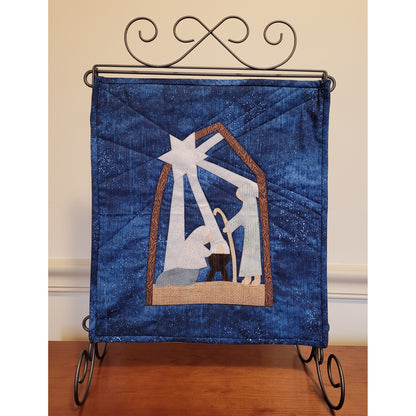 Festive quilt handing from metal stand. The quilt is the nativity scene with arch holding up the star to the top left, which is shining down with its beams on the scene below. Below is kneeling Mary, baby Jesus in manger, and standing shepherd with staff. Entire design on blue background fabric.