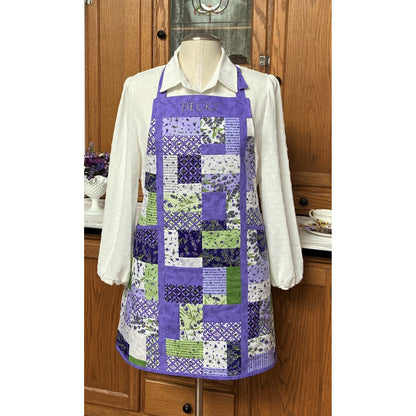 Pretty apron showcased on a mannequin with a white blouse underneath. Apron is made from patches of purple, green, and white fabric with piping of purple including top with person's name embroidered.