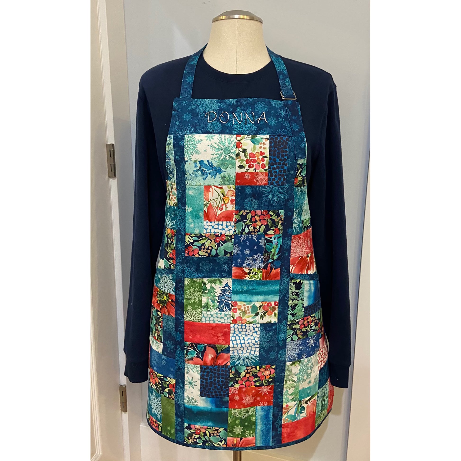 Pretty apron showcased on a mannequin with a dark sweatshirt underneath. Apron is made from patches of blue, red and green fabric with piping of blue/teal fabric including top with person's name embroidered.