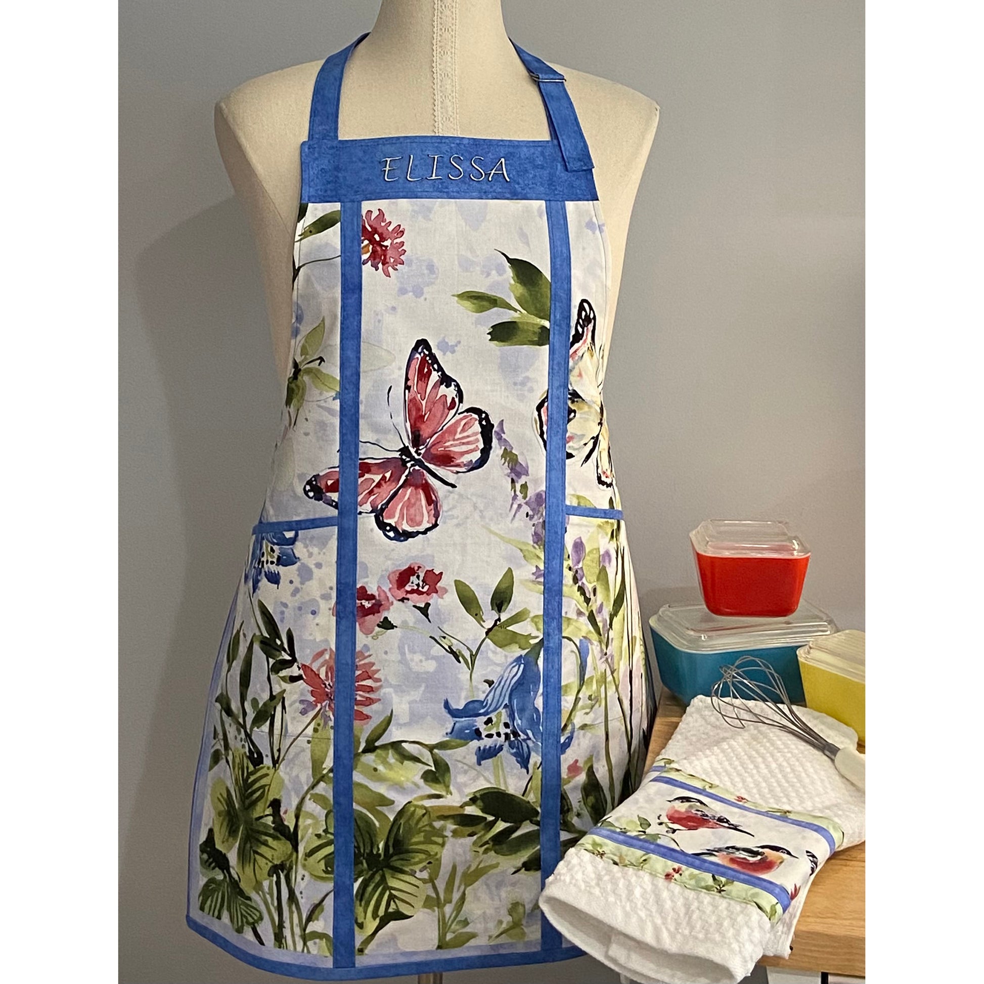 Beautiful apron shown on a mannequin with baking items near by. The apron is made of a panel with flowers and a butterfly. It is split on the sides with cording and has two large pockets on either side and the name of the owner on the top. 