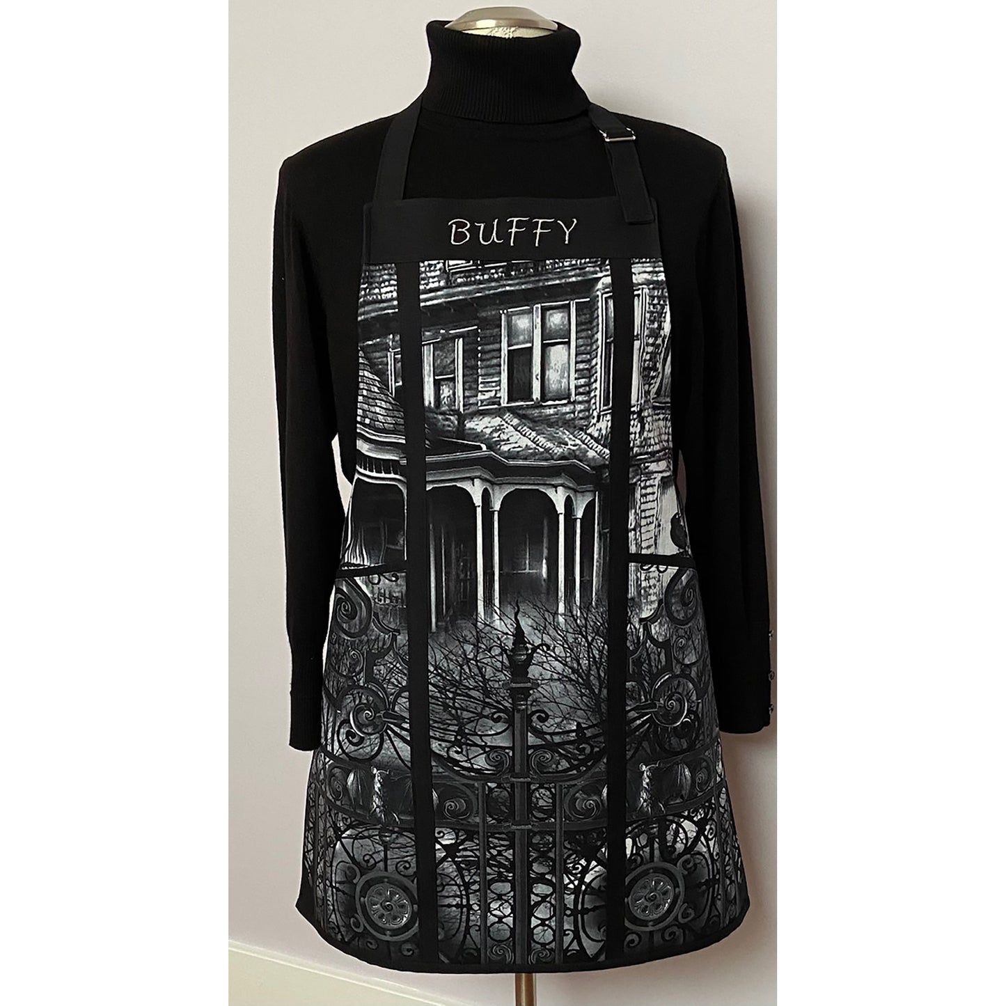 Beautiful apron is made of a panel in black and white of a haunted house. It is split on the sides with cording and has two large pockets on either side and the name of the owner on the top. 