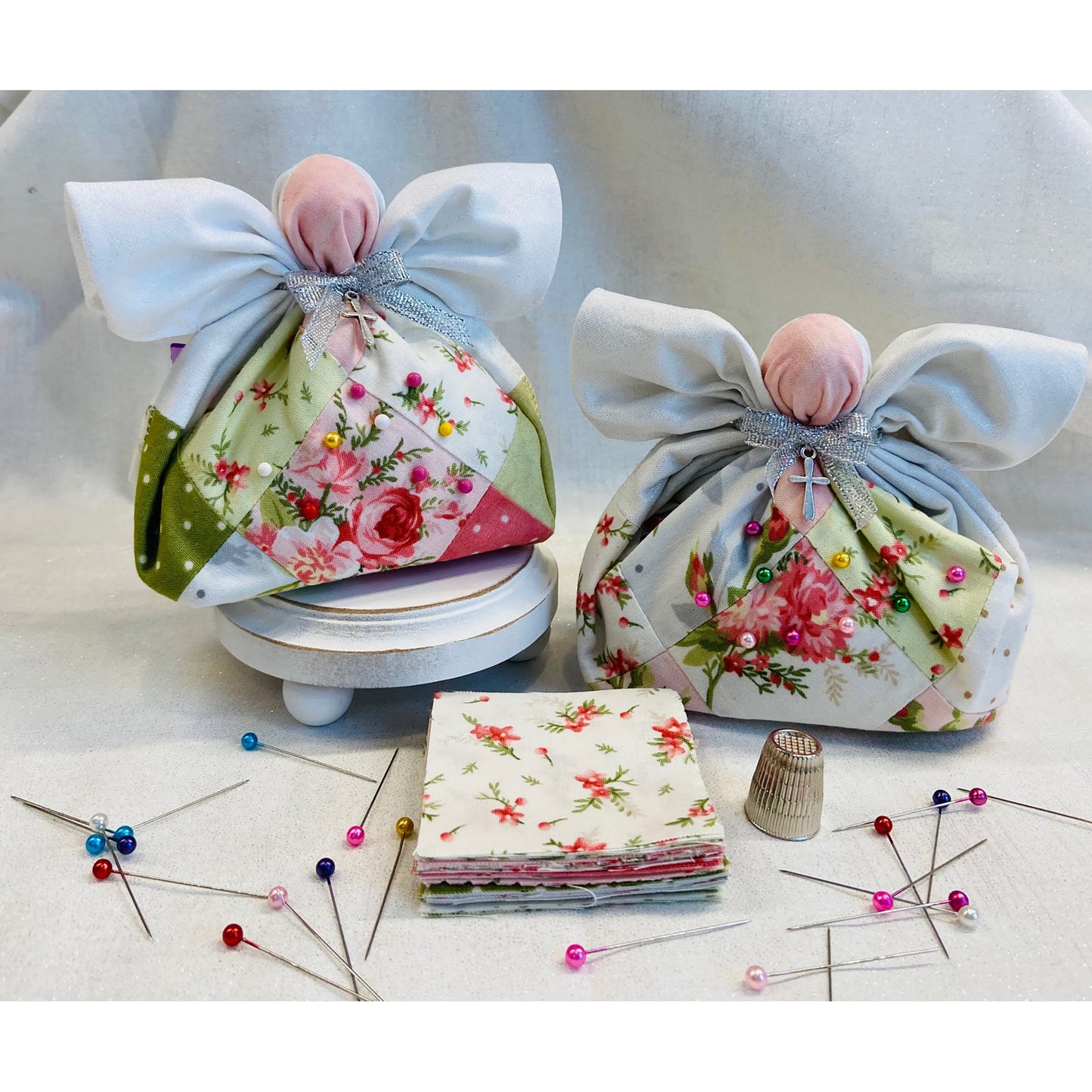 Two adorable pack work angel pincushions with pins to show how they work. Their patch work bodies hold pins. Around their necks are ribbons with a cross handing down. Thimble, pins and a small pile of fabric are shown in front.