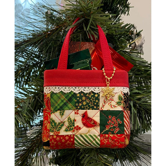 Small quilted tote bag with small candies inside. Hanging from a tree branch. Hanging from the one side of the tote handle is a jump ring with a snowflake charm.