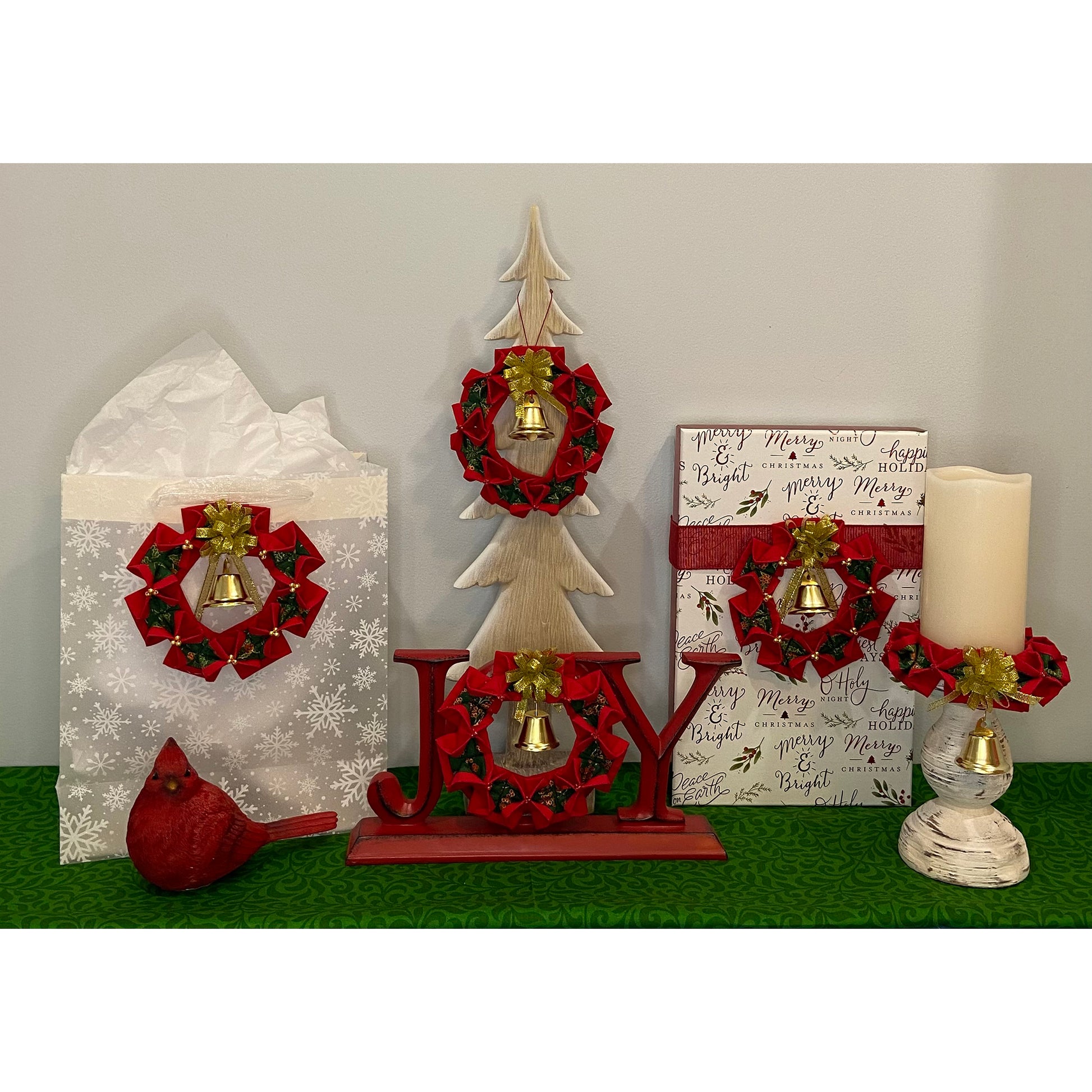 Seasonal display of ways to use the completed Christmas Wreaths ornaments. Shown as decoration on a gift bag, on a small wooden tree, in the O of Joy, on a gift box and around the base of a candle.