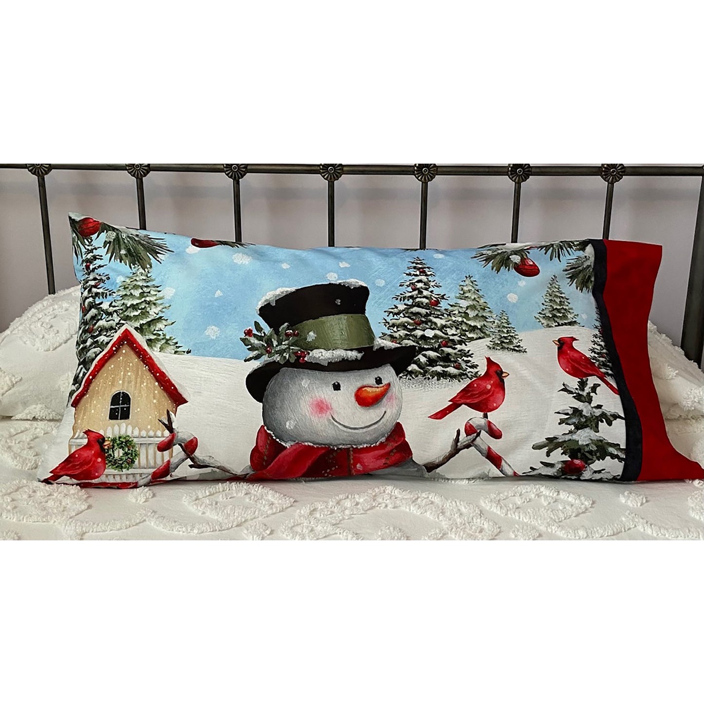 Pillowcase on a bed with a cute snowman in the snow holding a cardinal with a birdhouse next to him. Lots of snow covered pine trees in the background.