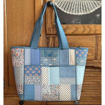 Beautiful tote bag hanging off a door in shades of blue fabric in patched blocks with light blue handle and piping around edges and two large pockets in middle.