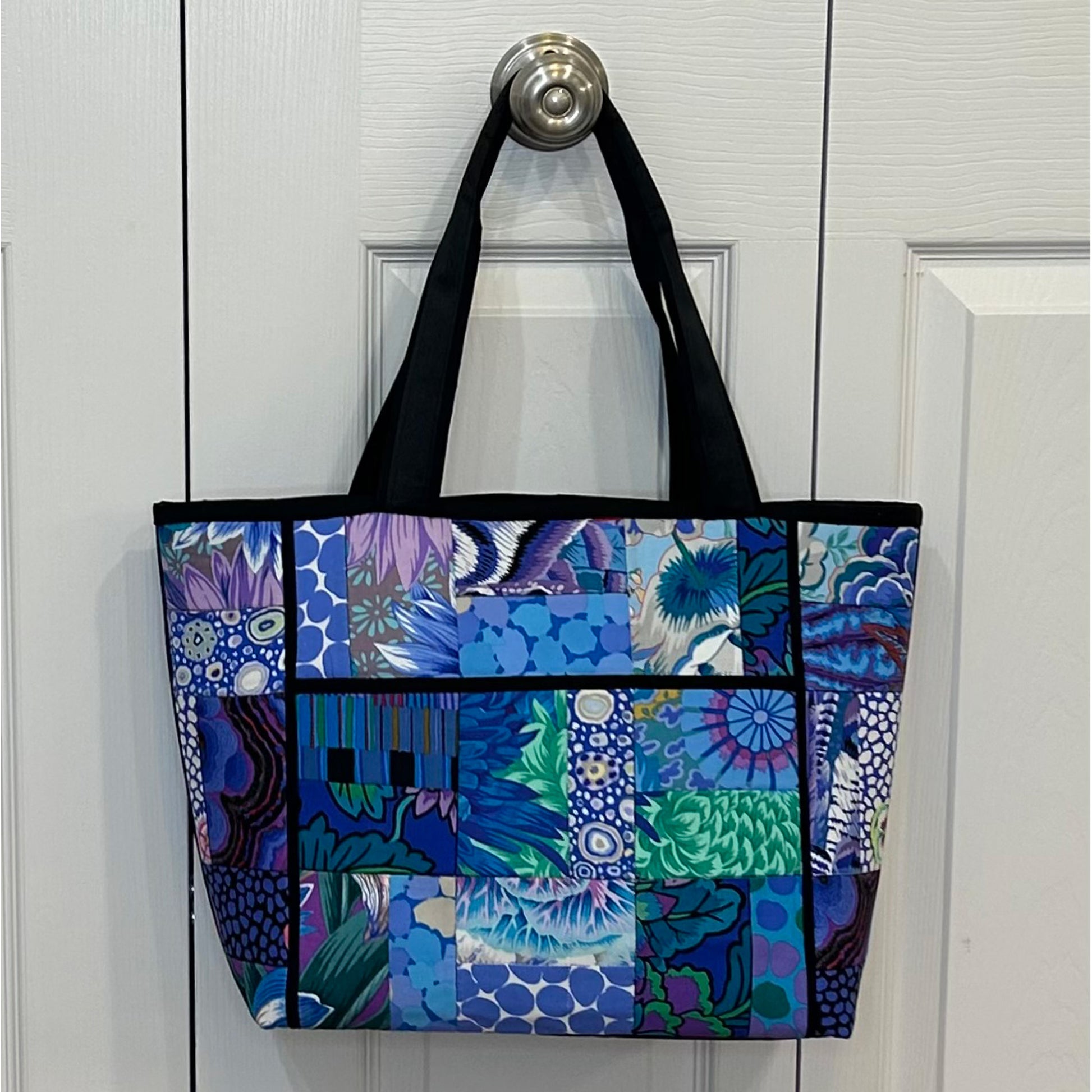 Classy floral tote bag in blue with contrasting colors floral fabric in patched blocks with dark handle and piping around edges and zipper pocket in middle.