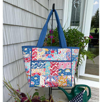 Beautiful tote bag hanging from a hook outside front door. Beautiful floral print fabric used in patched blocks with blue handle and piping around edges and zipper pocket in middle.