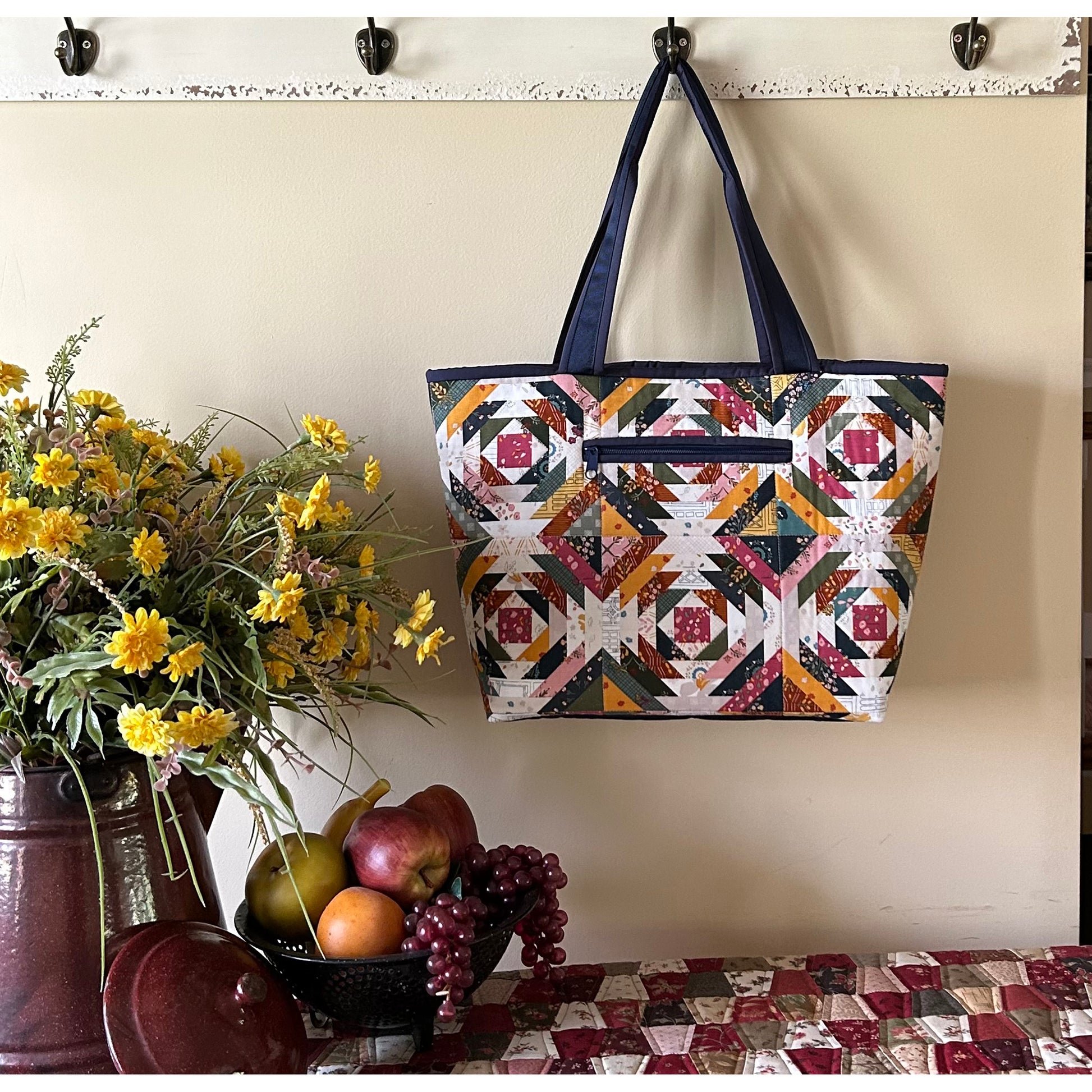 Beautiful tote bag hanging from a hook in the kitchen. Bag features log cabin fabric in bright colors with a zippered pocket in the middle.