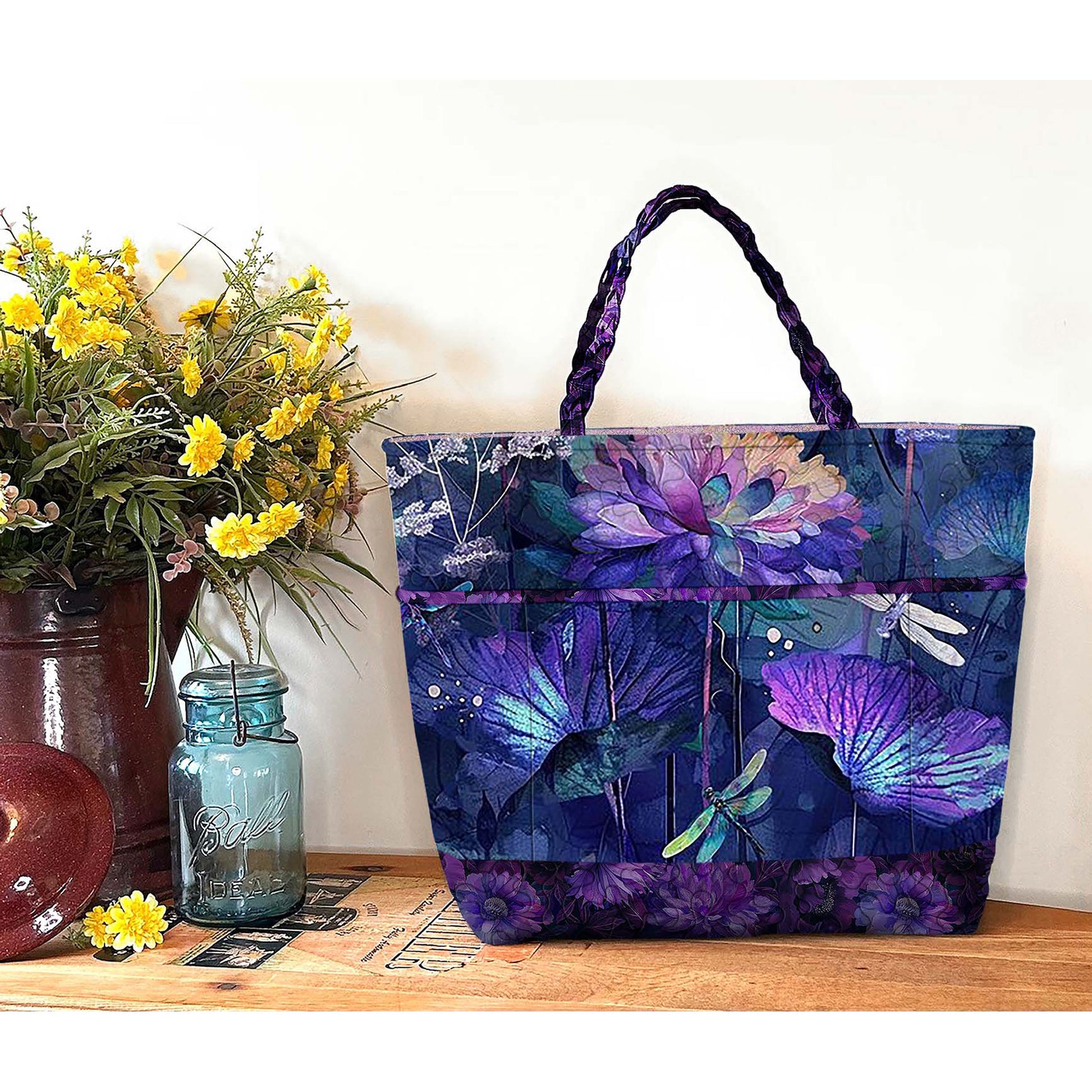 Tote bag with floral and dragonfly design on a wooden surface with flowers and a jar.