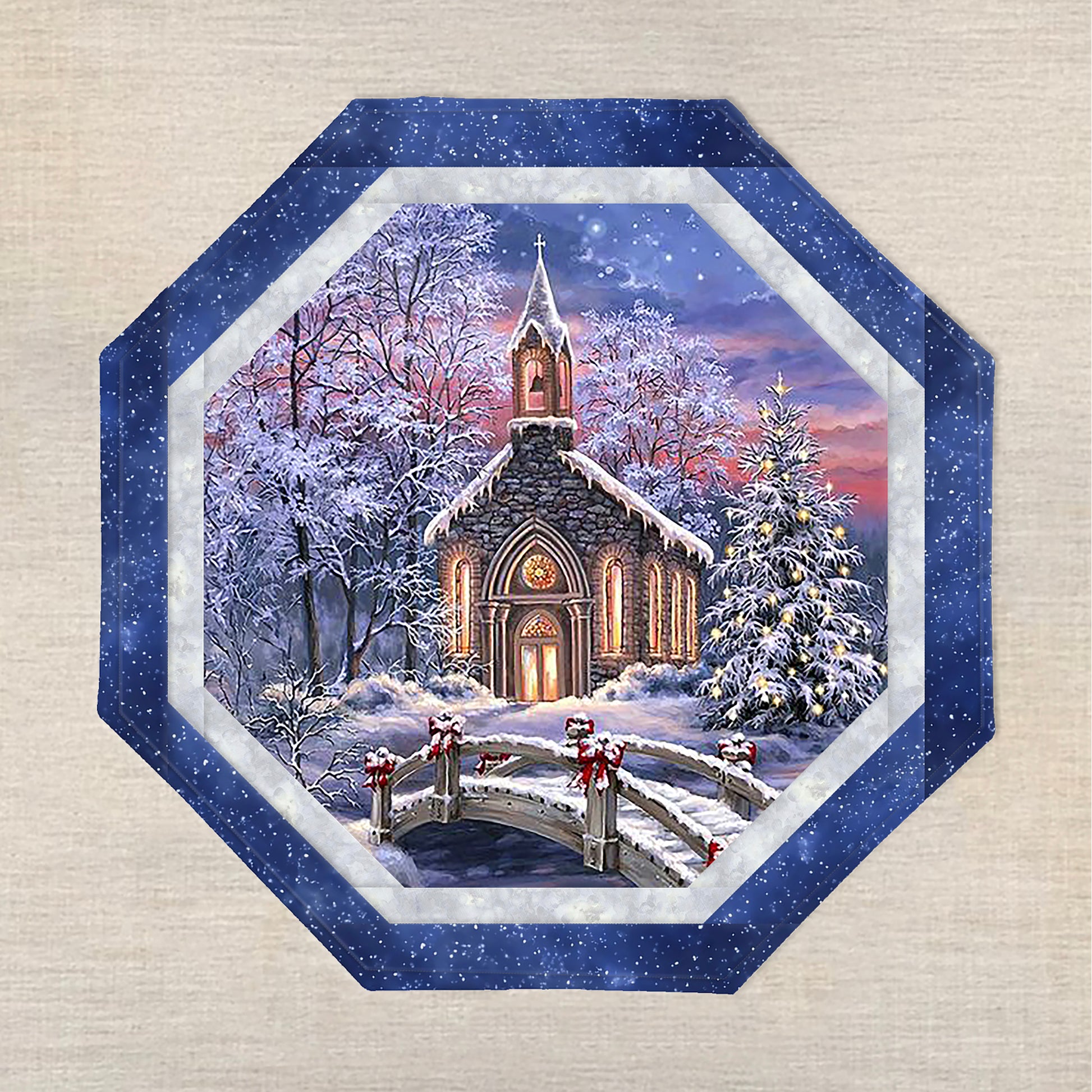 Octagon table topper features a print with snow covered church with a bridge and trees covered in snow. One tree is covered in white lights and the church is inviting with its lights on.