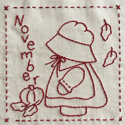 Sunbonnet Sue redwork embroidery design for November featuring Sunbonnet Sue in her pilgrim outfit picking a pumpkin as leaves fall.