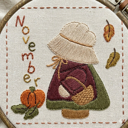 Sunbonnet Sue embroidery design for November featuring Sunbonnet Sue in her pilgrim outfit picking a pumpkin as leaves fall.