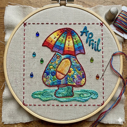Sunbonnet Sue colorful embroidery design for April featuring rain drops, boots and an umbrella hat.