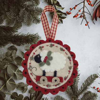 One ornament close up showcases a cream background and red scalloped edge with checker red and cream ribbon for hanging. The design on the ornament is a sheep on wheels with some holly around its neck.