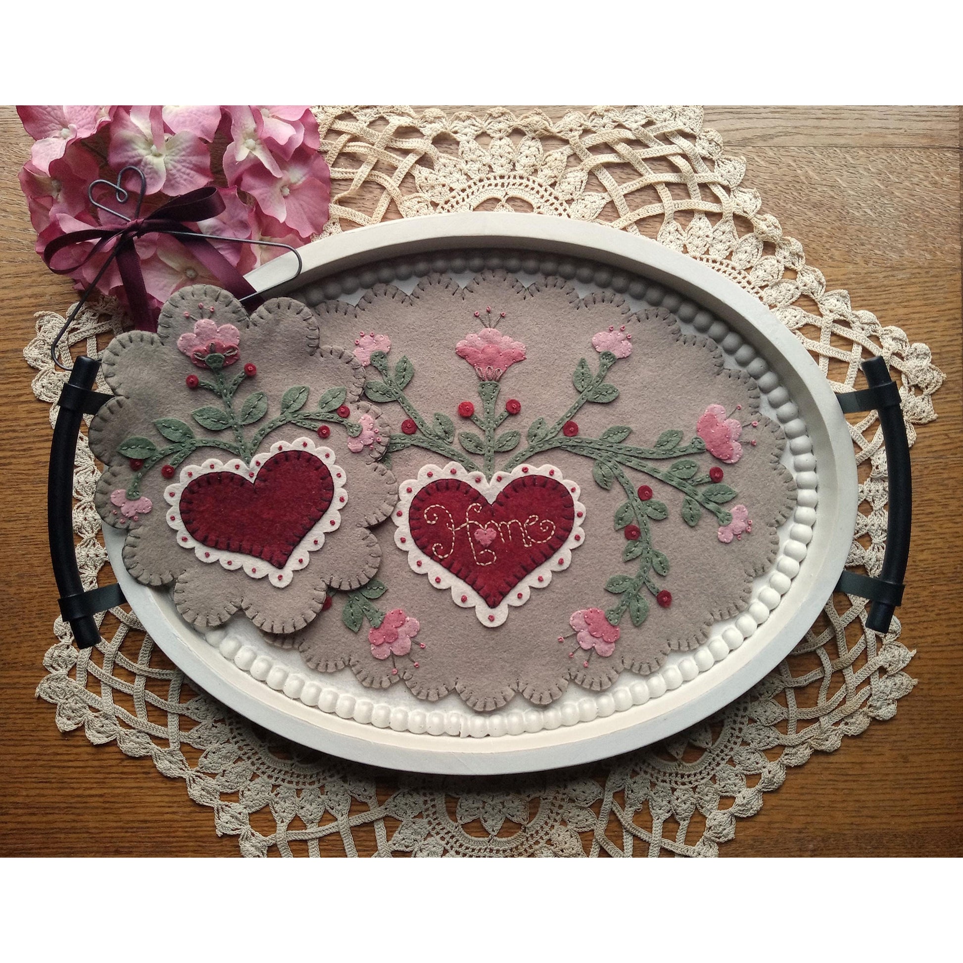 Two pretty wool felted mats. One is small on the left with a flower on top, scalloped edges and heart in the middle. The other takes up the middle of the image in a tray is oval and has a heart with the words "home" inside it and flowers all around it. Also scalloped edges.