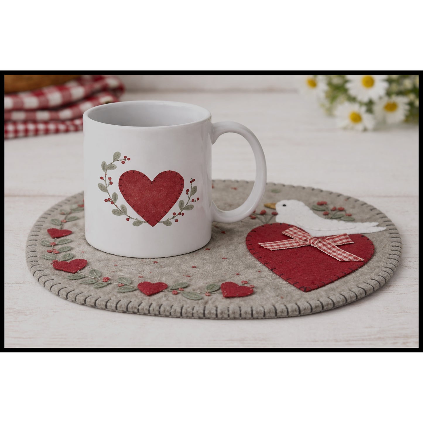 Pretty mug rug in wool felt features a heart with a dove in it and hearts border. This image includes a matching heart mug on top.