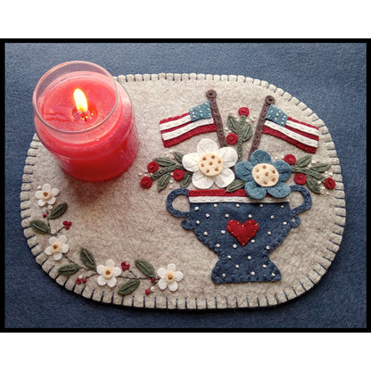 Wool candle mat is a heirloom-style pattern features a lovely vase filled with pretty flowers and two American flags.