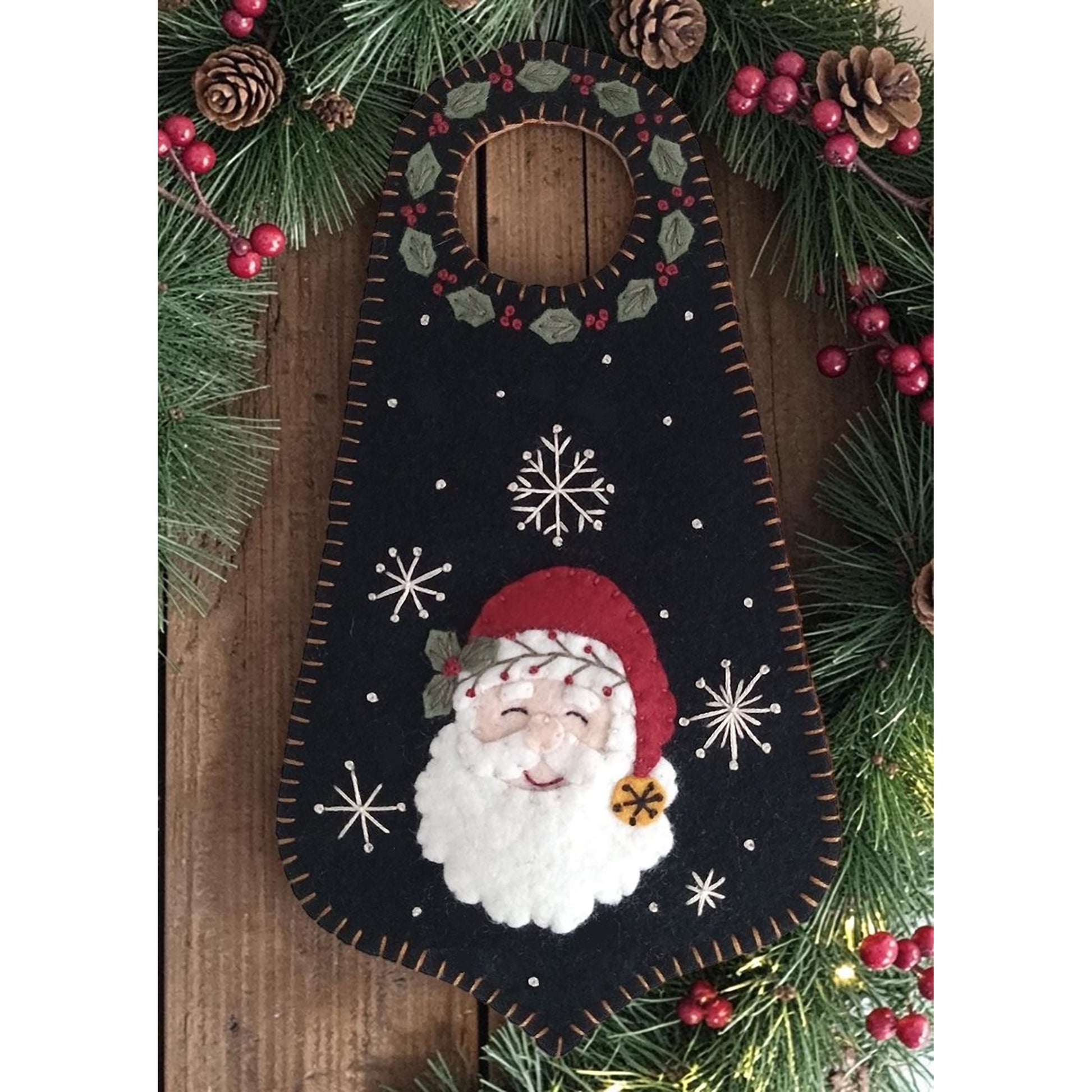 This sweet wool door hanger features a smiling Santa surrounded by winter greenery, berries, and pretty snowflakes. 
