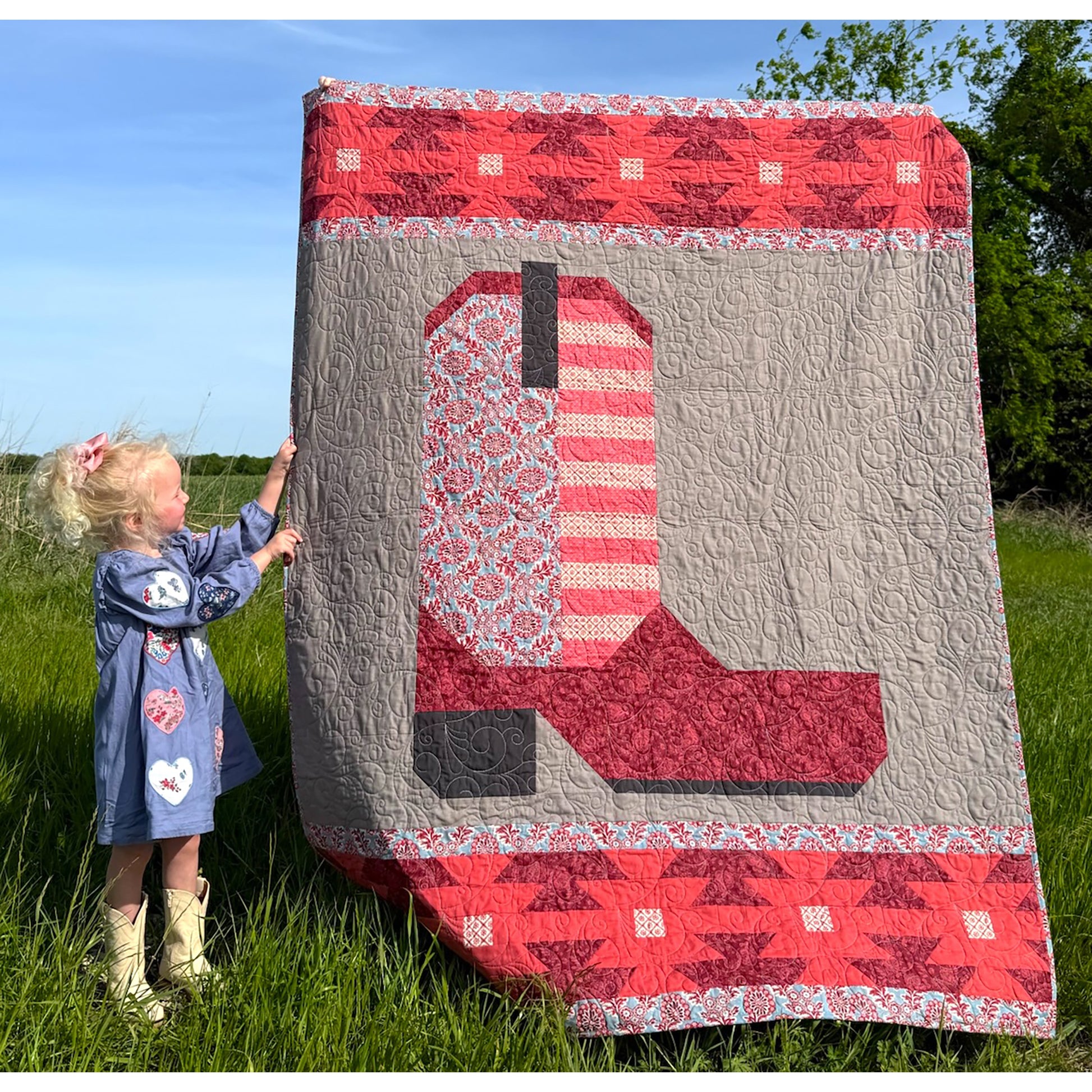The Heartland Boot quilt is a reflection of our roots that are deep in the Heartland of America. Designed for all the farmers, ranchers and a life tied directly to the land. This quilt is in shades of pink with a young girl standing next to it.