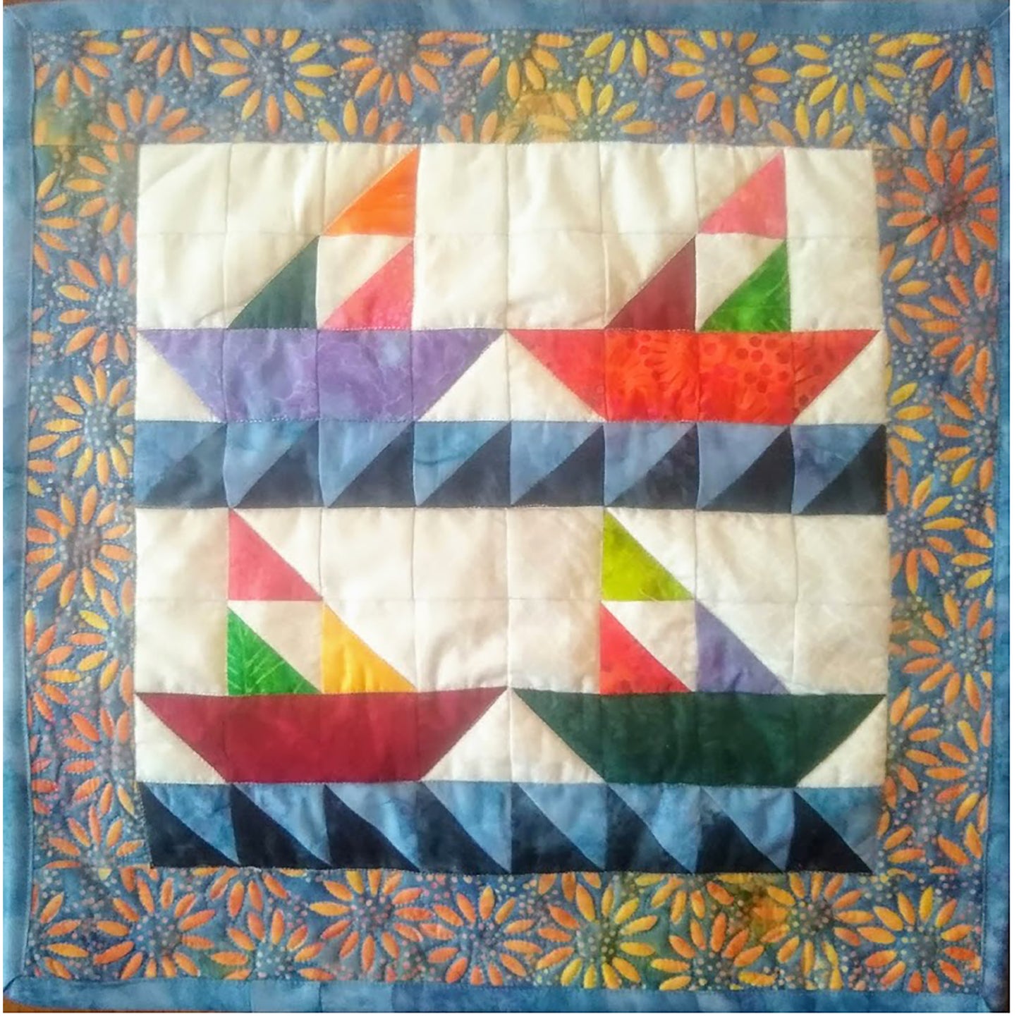 Four pieced sail boats made from bright colored fabrics with blue waves. The top two appear to be heading to the right and bottom two to the left. Their sails are made from three triangles.